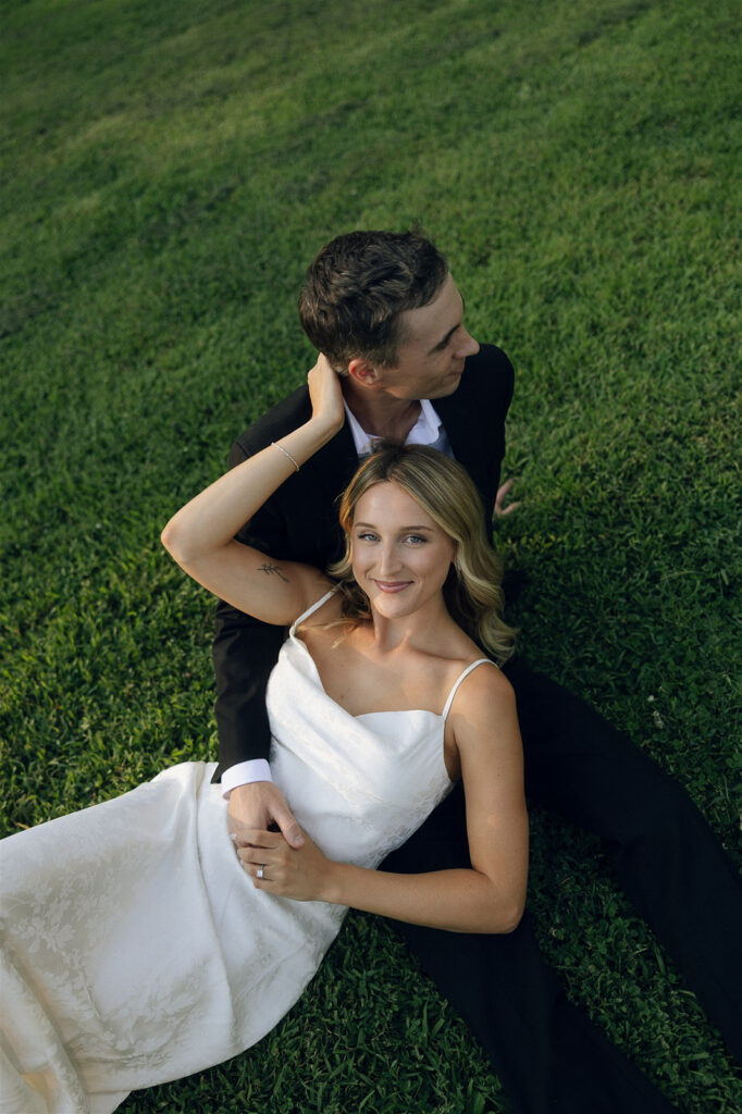 Engaged couple lying in green grass at golden hour, she looks up at the camera while he wraps his arms around her during their romantic St. Paul engagement photos.