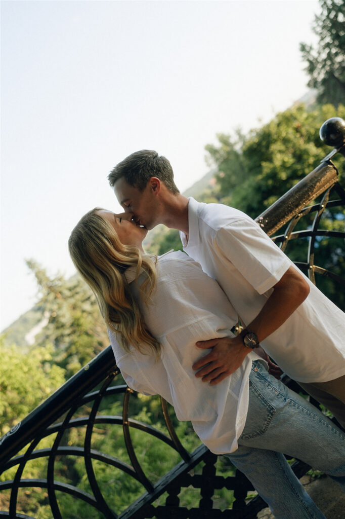 He dips her slightly against the black iron railing and kisses her, one hand wrapped around her waist, green hills and soft summer light stretching out behind them.