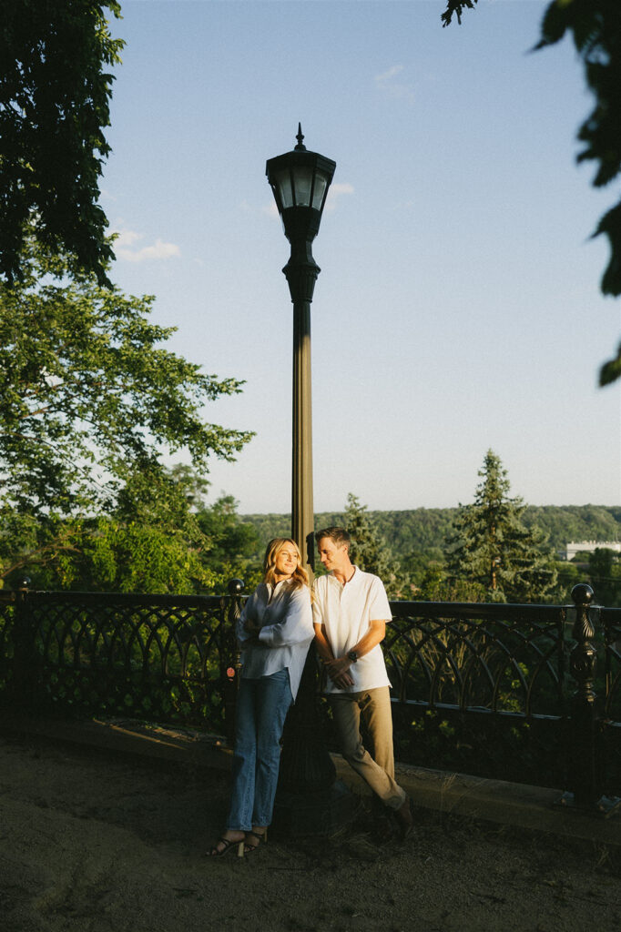 They stand beneath a tall lamppost at golden hour, leaning casually against the railing and smiling at each other with the overlook and tree-covered hills in the distance.