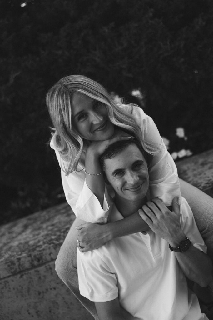 Black and white portrait of her perched on his back, arms wrapped around his shoulders, both of them smiling softly toward the camera.
