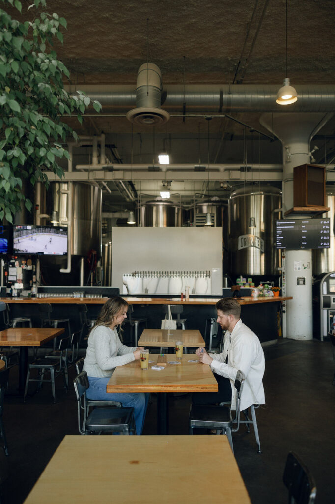 Wide view of the taproom as they sit across from each other playing cards, the kind of engagement photos unique to people who actually like hanging out.