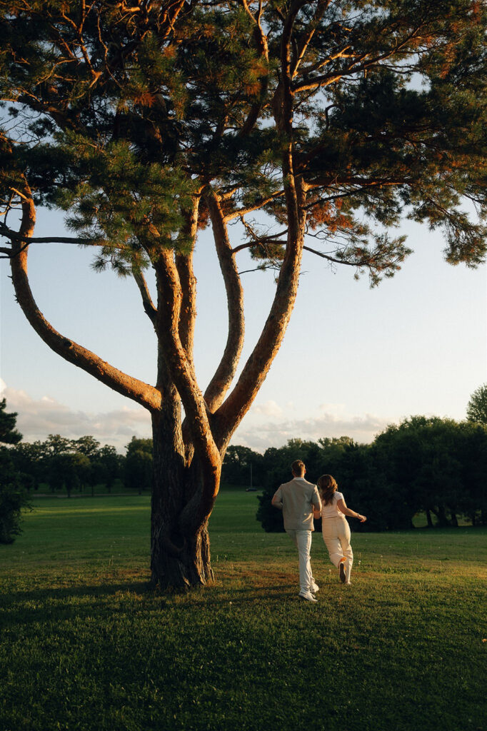 Wide golden hour engagement photo of a couple running hand in hand beneath a towering tree in a Minneapolis park, late summer light stretching long shadows across the grass.