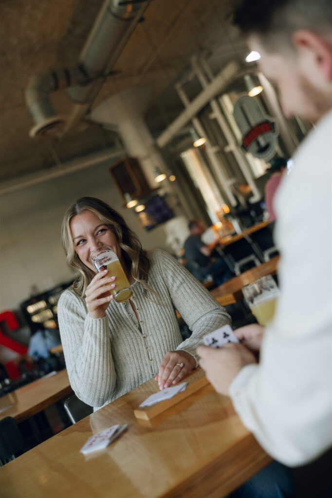 She laughs mid-sip across the table, cards scattered and date-night energy fully intact — engagement photos that feel like a Tuesday night, not a production.