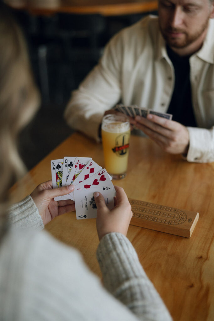 Over-the-shoulder shot of a couple playing cards at a Minneapolis brewery, pints sweating on the table between them.