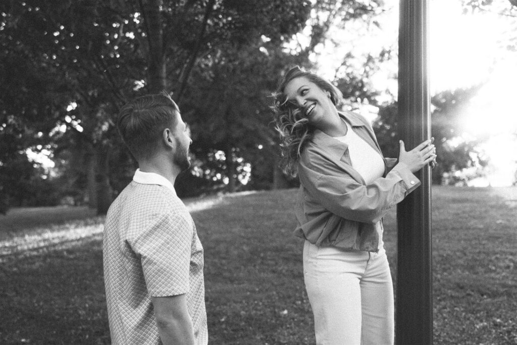 Black and white engagement photo of a couple at golden hour in a Minneapolis park, she’s mid-laugh holding onto a lamppost while he looks at her like he already knows she’s about to cause trouble.