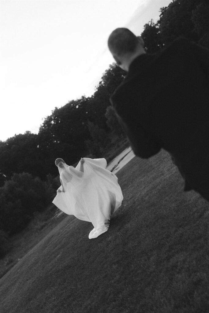 Black and white photo of bride walking through vineyard lawn at 7 Vines Vineyard and Winery, groom following behind as her veil lifts in motion.
