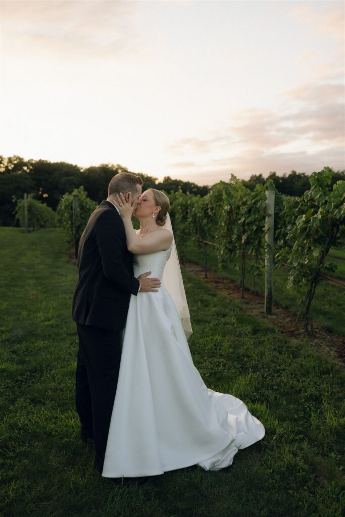 Bride and groom kissing in the vineyard at golden hour, soft August light glowing over 7 Vines Vineyard and Winery.
