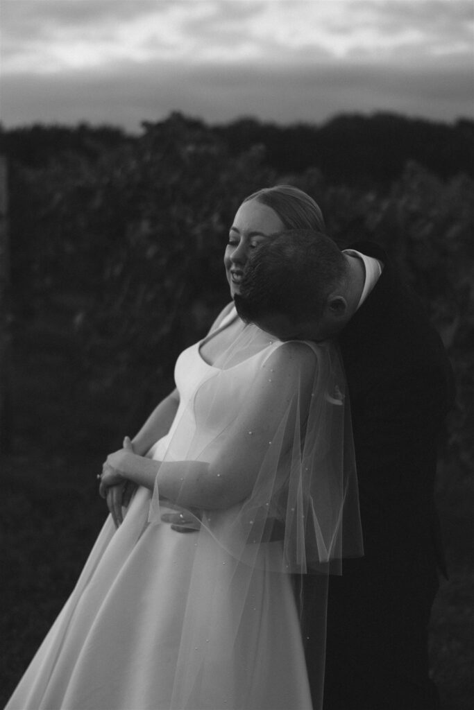 Romantic black and white portrait of bride leaning into groom among vineyard rows at 7 Vines Vineyard and Winery.