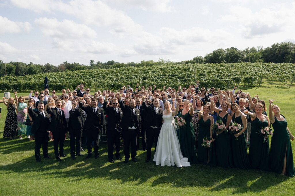 Large group champagne toast photo at 7 Vines Vineyard and Winery, wedding guests raising glasses in front of the vineyard on a vibrant summer afternoon.