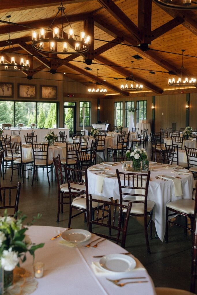Reception space at 7 Vines Vineyard and Winery set with round tables, white linens, gold flatware, and soft floral centerpieces beneath warm wood beams and modern chandeliers.