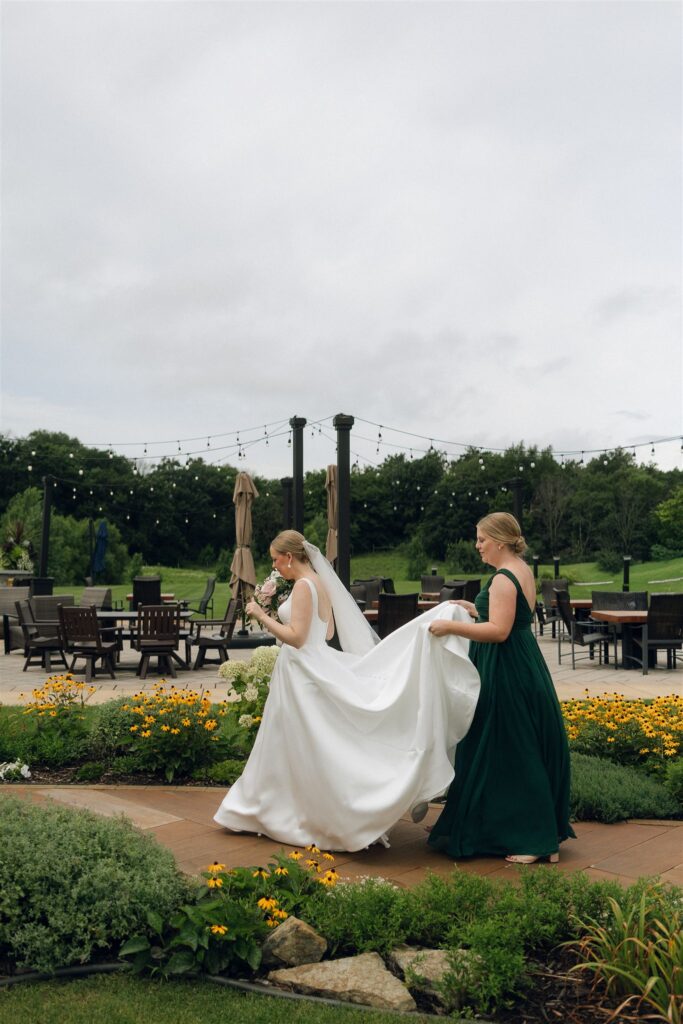 Bride walking across patio with bridesmaid adjusting train, vineyard and string lights in background at 7 Vines Vineyard and Winery.