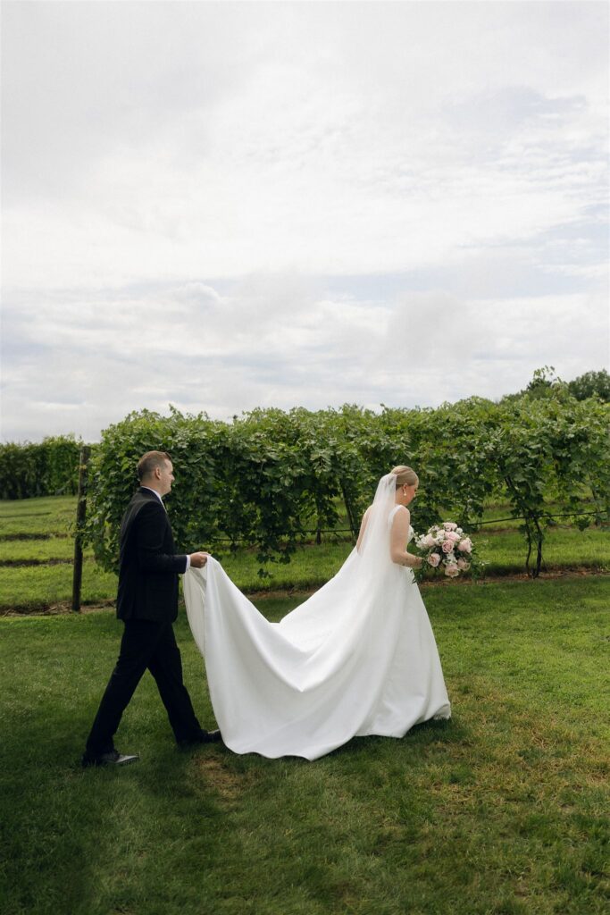 Bride walking through vineyard lawn with groom holding her train, summer greenery surrounding them at 7 Vines Vineyard and Winery.