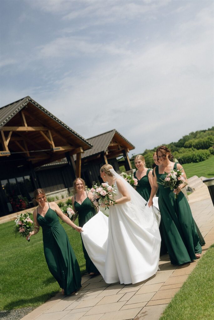 Bride and bridesmaids in deep green dresses walking along the patio at 7 Vines Vineyard and Winery under bright summer sun.