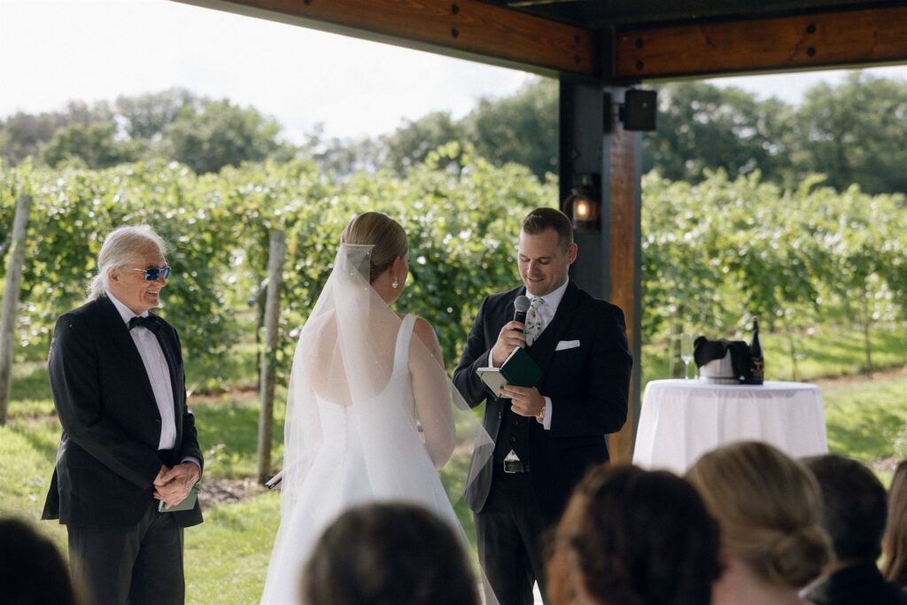 Groom reading vows during an outdoor ceremony at 7 Vines Vineyard and Winery, vineyard vines lush and green in the background.