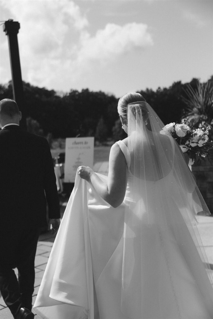 Bride walking toward the ceremony space in a pearl-detailed veil at 7 Vines Vineyard and Winery, summer light casting soft shadows.