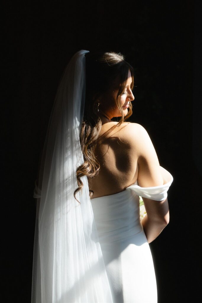 Bride in an off-the-shoulder gown standing in window light, her reflection visible in the glass door inside the vineyard reception space.