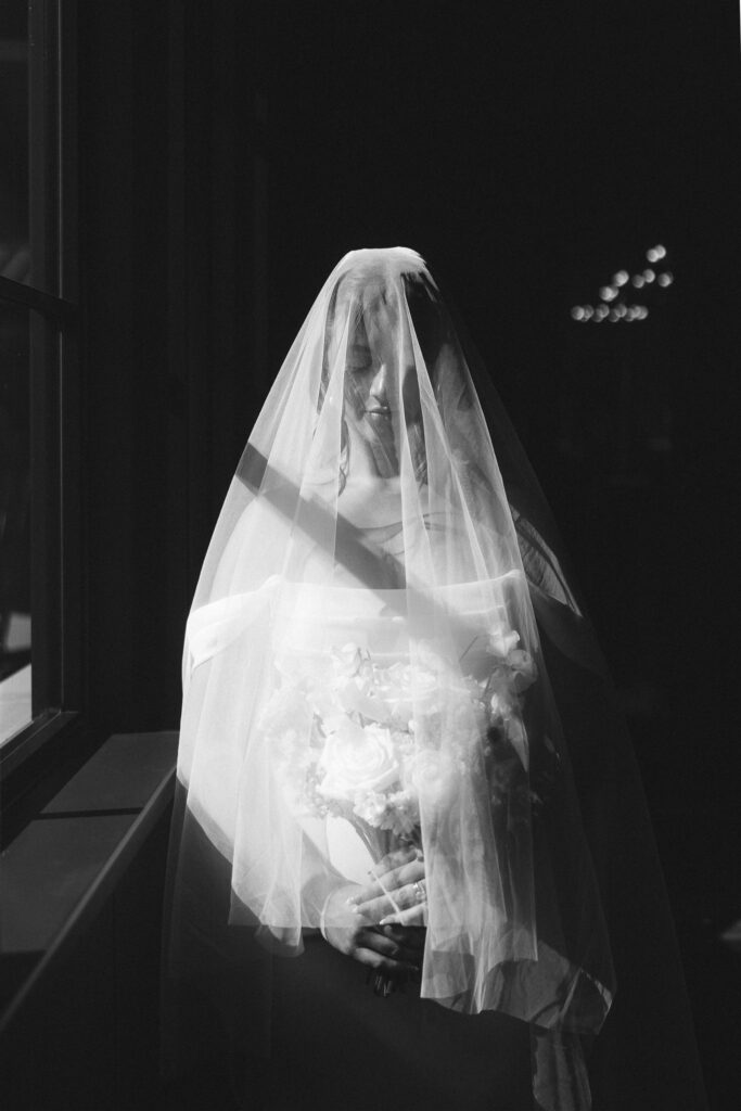 Black-and-white portrait of bride with veil draped over her face, bouquet of white roses softly visible beneath the tulle.