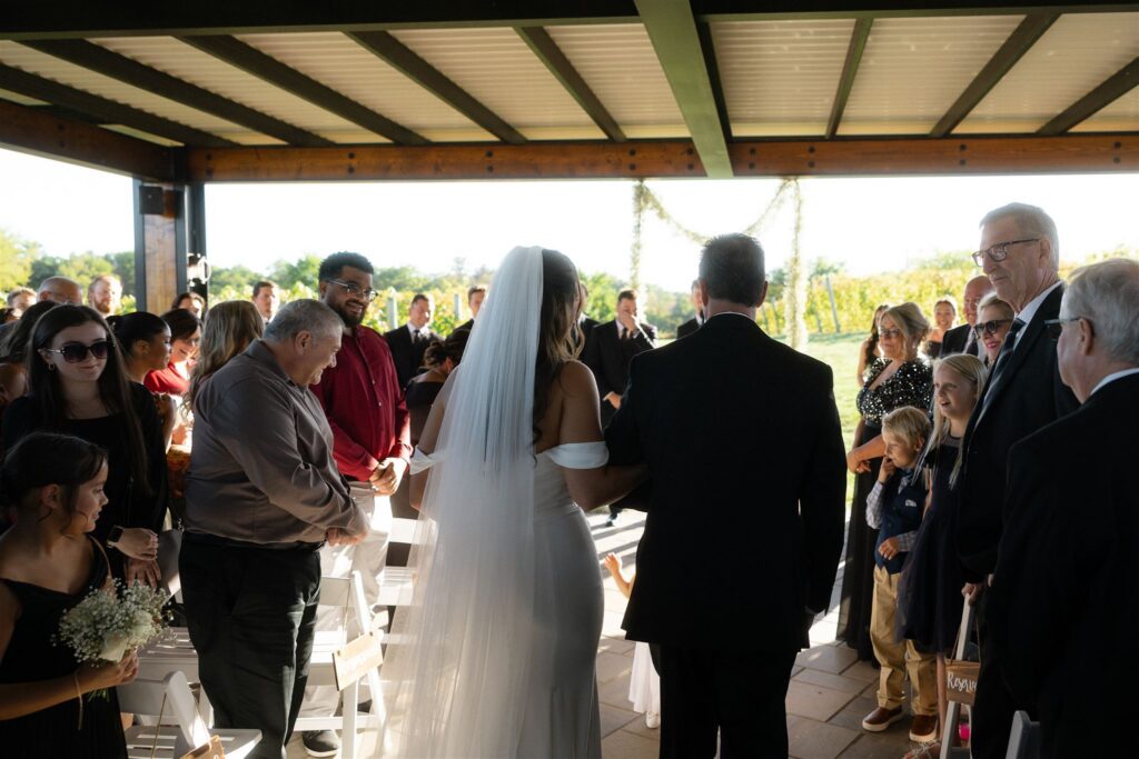 Bride walking down the aisle at 7 Vines Vineyard and Winery with guests standing on either side beneath the covered patio.