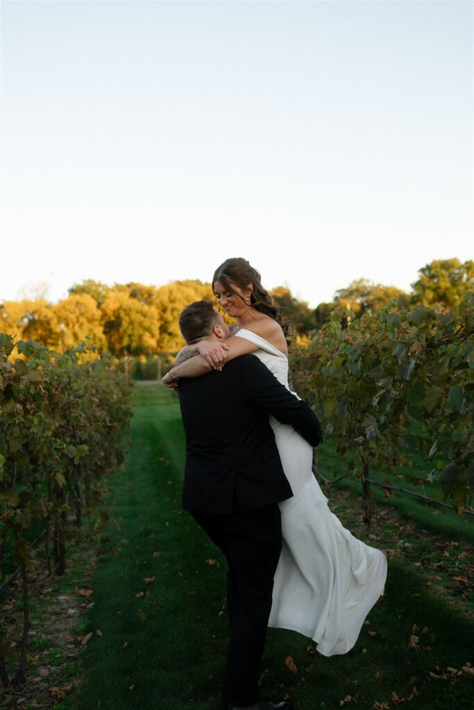 Bride and groom walking hand in hand through vineyard rows at 7 Vines Vineyard and Winery, early autumn leaves beginning to turn under golden hour light.