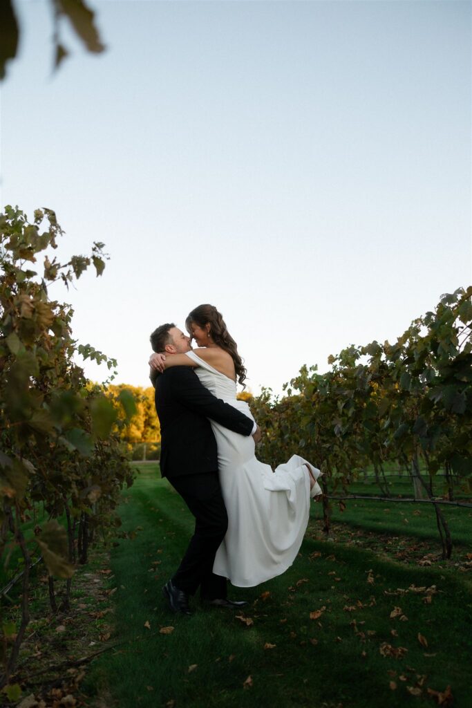 Groom lifting his bride in the vineyard at 7 Vines Vineyard and Winery, her off-the-shoulder gown flowing as golden October light filters through the rows of vines.