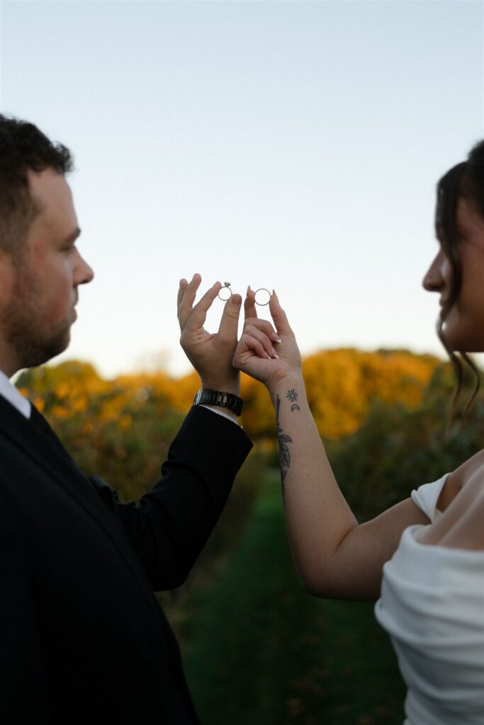 Close-up of bride and groom holding up their wedding rings between them in the vineyard at 7 Vines Vineyard and Winery, warm fall foliage softly blurred in the background.