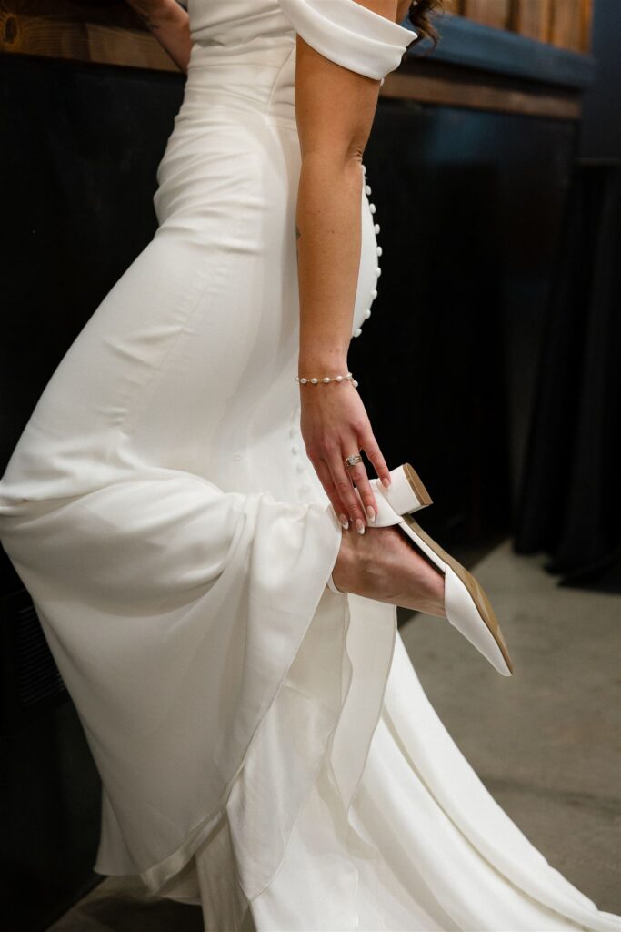 Close-up of a bride in an off-the-shoulder satin gown adjusting her white block heel, pearl bracelet and engagement ring visible.