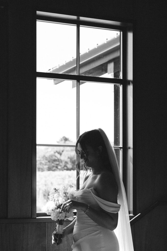 Black-and-white bridal portrait by the window at 7 Vines Vineyard and Winery, bride holding a white bouquet with soft light outlining her veil.