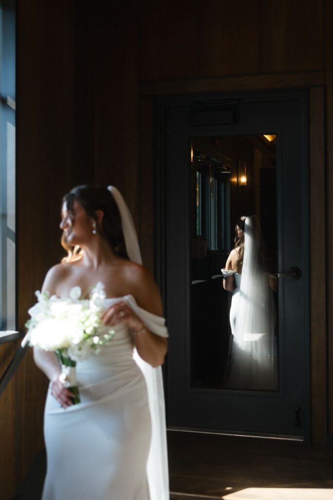 Bride in an off-the-shoulder gown standing in window light, her reflection visible in the glass door inside the vineyard reception space.