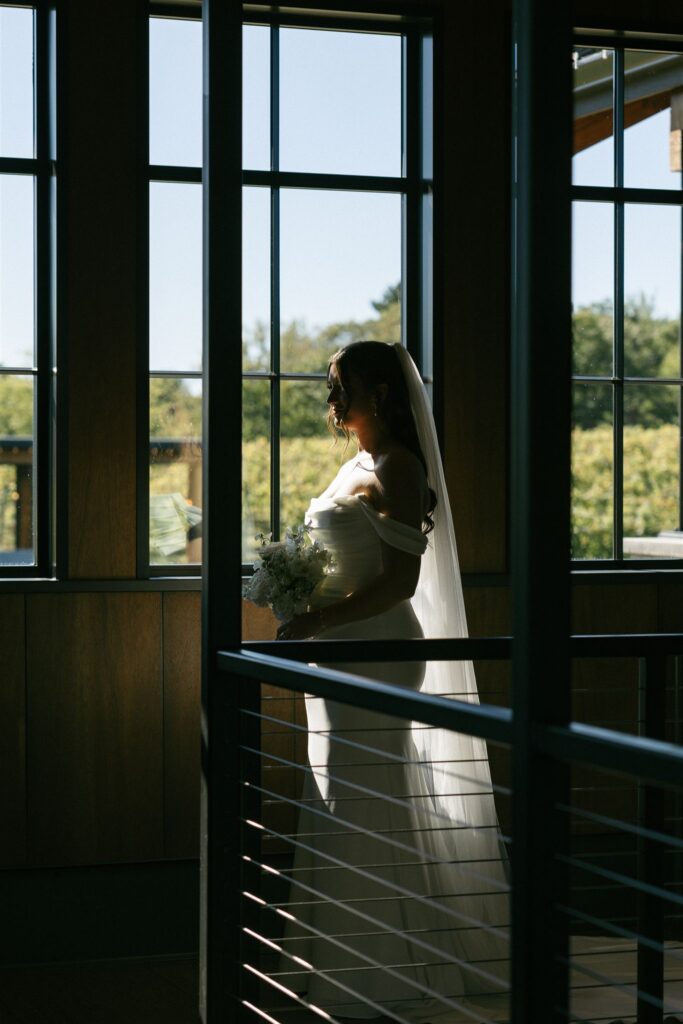 Silhouette of bride holding a bouquet in front of tall windows overlooking the vineyard rows at 7 Vines Vineyard and Winery.