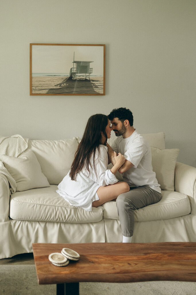 Couple cuddled close on a cream couch during an in-home engagement session, foreheads touching as they sit knee-to-knee, soft natural light filling the space