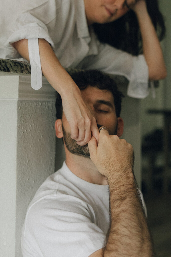 Man kissing his fiance's fingers as they reach towards him from where she lays on the countertop. 