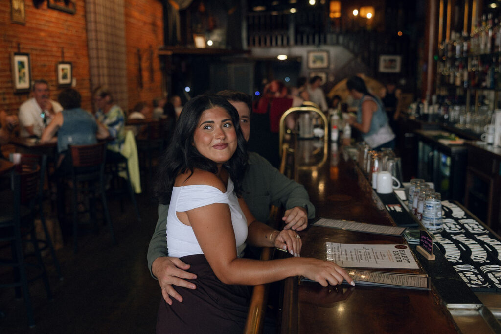 Couple sitting at a dimly lit bar during their engagement session, his arm wrapped around her waist as she leans back into him—an engagement photo idea rooted in physical touch and effortless closeness.
