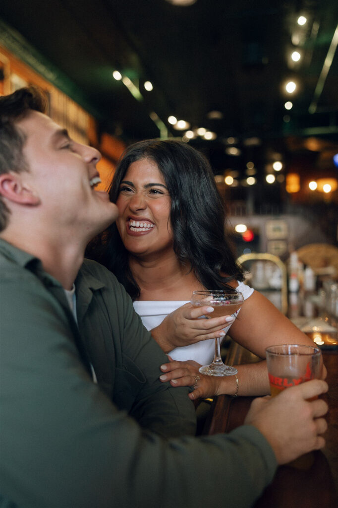 Close-up engagement photo of a couple laughing mid-conversation at a bar, her hand resting on his arm while they lean in close, showcasing candid physical touch.