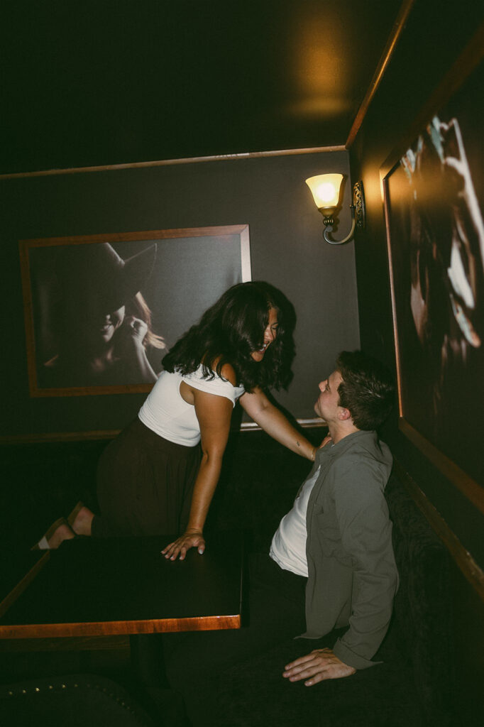 Moody engagement photo of a couple tucked into a booth, she kneels on the seat as he looks up at her, bodies drawn together in a playful, cinematic moment.