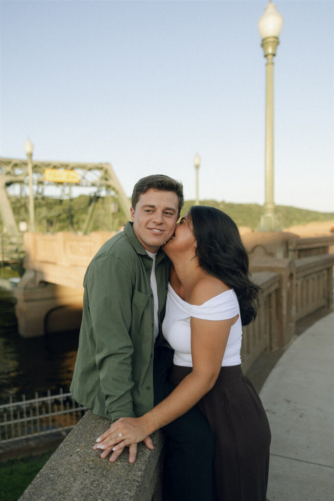 Engagement photo of a couple kissing on a bridge at golden hour, her hand resting on his shoulder while he leans into the moment, highlighting physical touch in an outdoor setting.