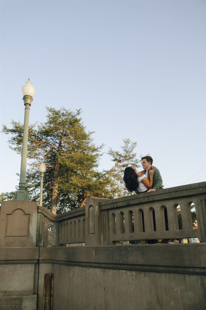 Wide engagement photo of a couple embracing on a bridge, surrounded by soft evening light and open sky, emphasizing closeness through simple body language.