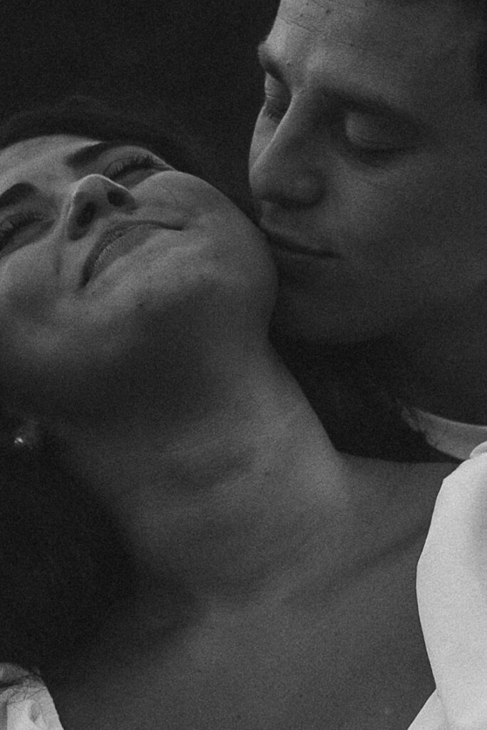 Grainy black and white close-up engagement photo of a couple cuddled together, his face pressed into her neck as she relaxes into the moment, highlighting quiet physical touch.