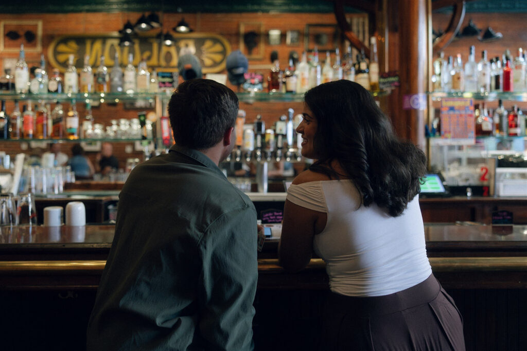 Couple leaning against a bar during their engagement session, bodies angled toward each other as they talk and smile, an engagement photo idea built around closeness and low-key physical touch.