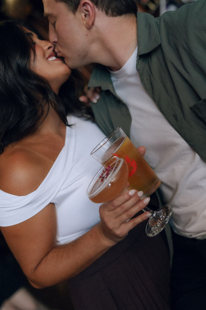 Close-up engagement photo of a couple kissing while holding drinks at a bar, her hand tucked into his shoulder as they lean in without hesitation, capturing physical touch in a candid moment.