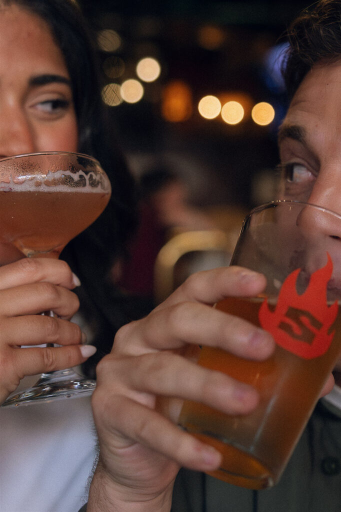 Tight engagement photo of a couple mid-sip, exchanging knowing looks over their drinks with warm bar lights blurred behind them, a playful engagement photo idea rooted in connection and chemistry.