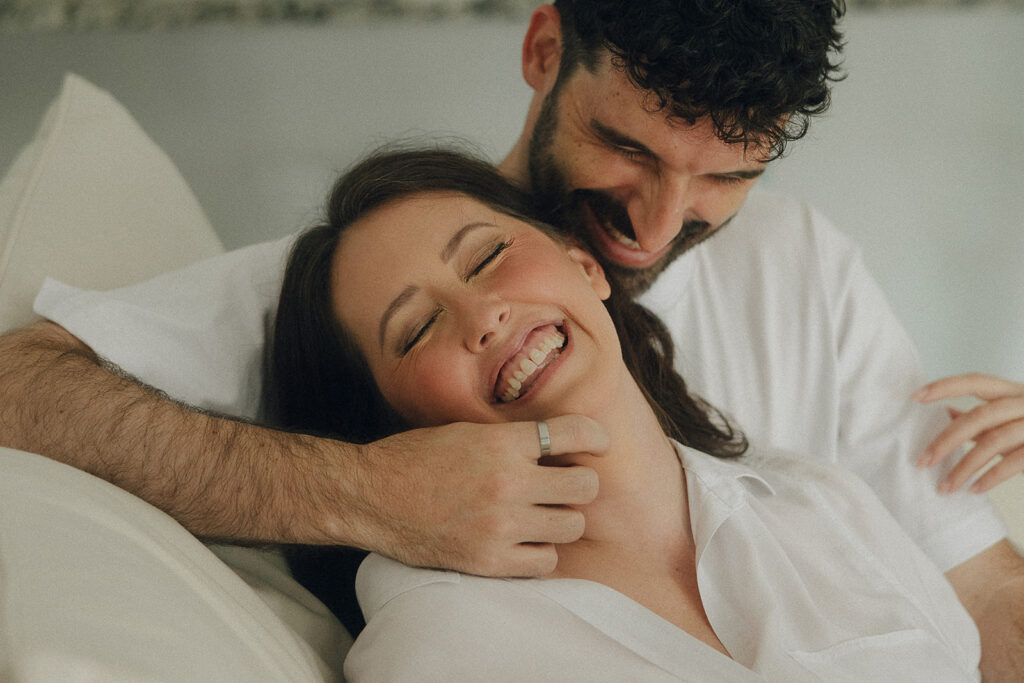 Playful in-home engagement photo of a couple laughing as he wraps his arm around her shoulders, physical touch showing up naturally without instruction