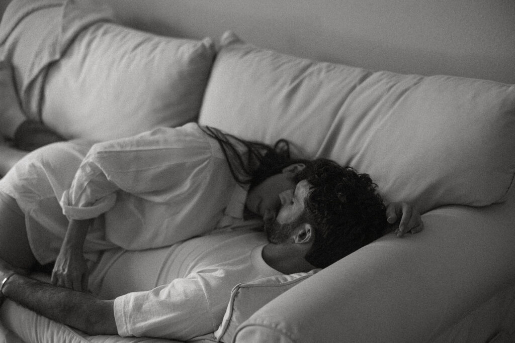 Lifestyle engagement photo of a couple stretched out on the couch, bodies overlapping and relaxed, capturing the quiet comfort of being at home together