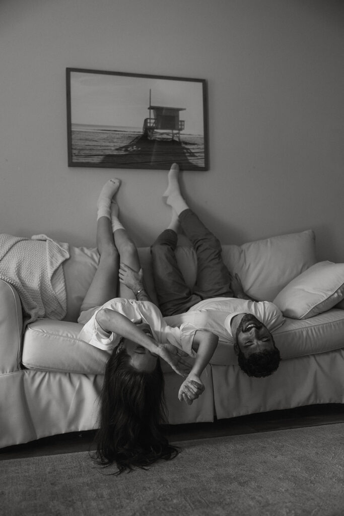 Black and white engagement photo of a couple upside down on the couch, hands intertwined and laughing, showing connection through playful physical closeness