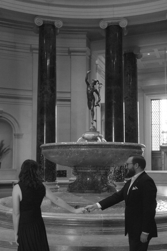Couple holding hands beneath a large classical fountain inside an art museum rotunda with tall columns.