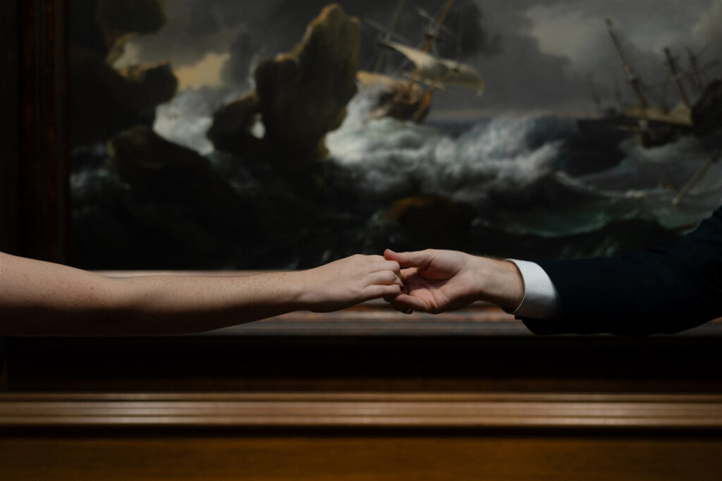 Close-up of two hands reaching toward each other in front of a dramatic classical painting.