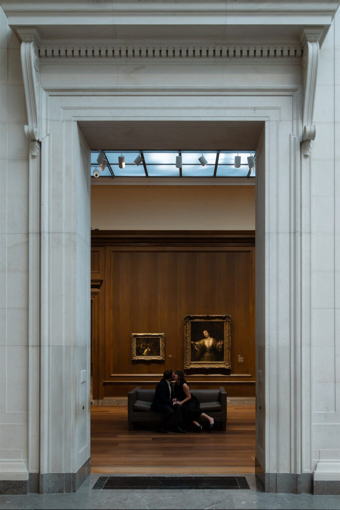 View through a white doorway into a museum gallery where a woman sits on a bench facing framed artwork.