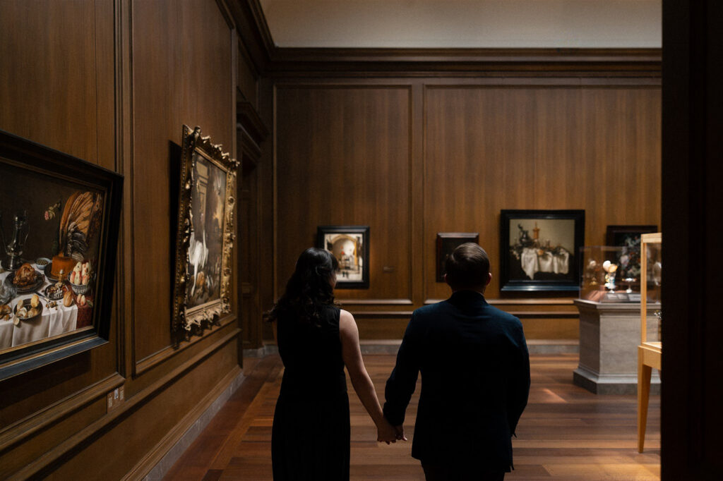 Couple walking hand-in-hand through a museum gallery lined with framed paintings and wood-paneled walls.