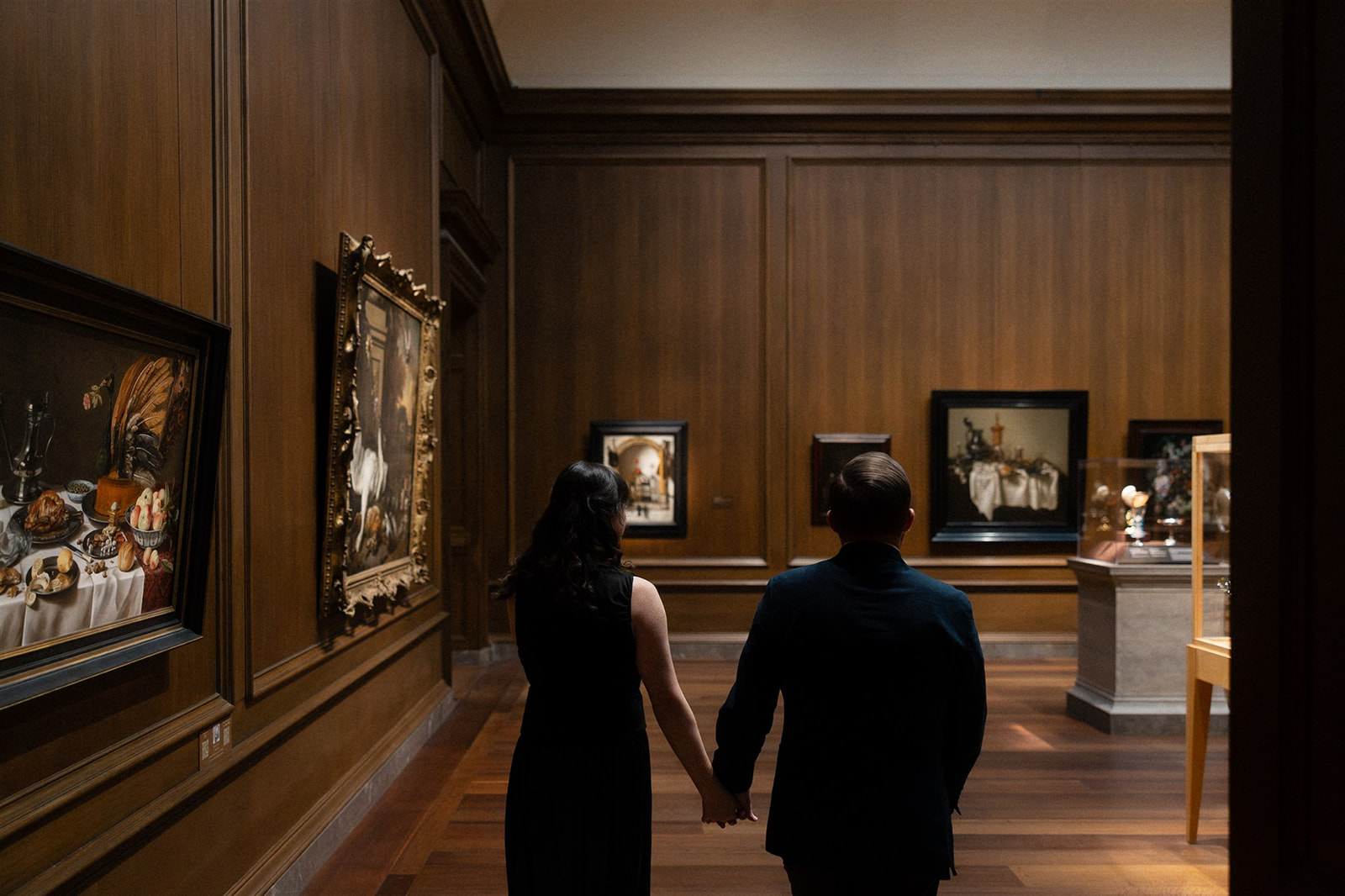 Back view of couple holding hands while walking through a museum gallery lined with classical paintings — cinematic engagement photography in an art museum setting.