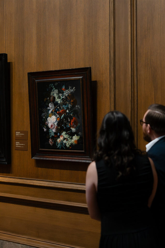 Couple standing side-by-side viewing a framed floral still-life painting in a museum gallery.