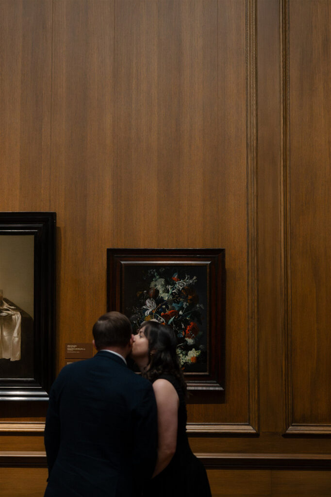 Couple leaning in closely together while looking at framed artwork on a wood-paneled museum wall.
